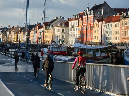 This urban photograph captures cyclists riding along a dedicated bike path in Copenhagen during the afternoon in autumn. The main subject of the image is Copenhagen cycling, set against a backdrop of the city’s iconic architecture. Distinctive multicoloured historic buildings line the waterfront in the Nyhavn district, a well-known landmark in Copenhagen. Boats are moored along the canal, and the sunlight casts a warm glow over the city, emphasizing the vibrant architectural style typical of Copenhagen. The image effectively showcases cycling as an integral part of urban life in this city, while highlighting the architectural heritage and waterfront scenery unique to Copenhagen.